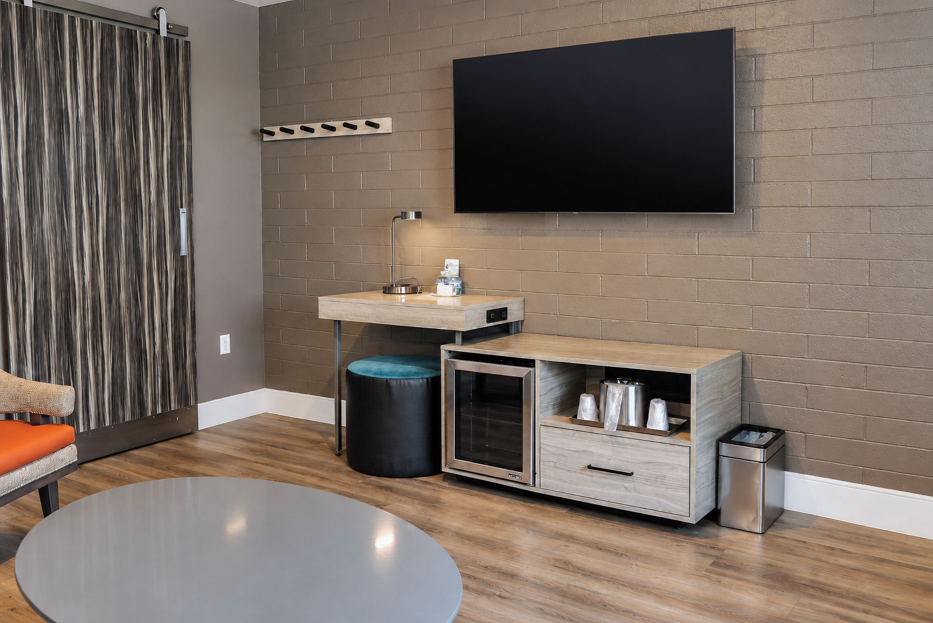 A modern room with a flat-screen TV mounted on a brick wall, a wooden vanity with a sink, a small trash bin, and a round table.
