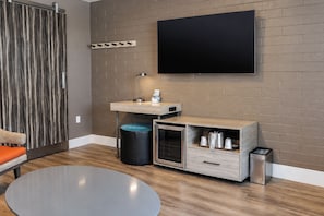 A modern room with a flat-screen TV mounted on a brick wall, a wooden vanity with a sink, a small trash bin, and a round table.
