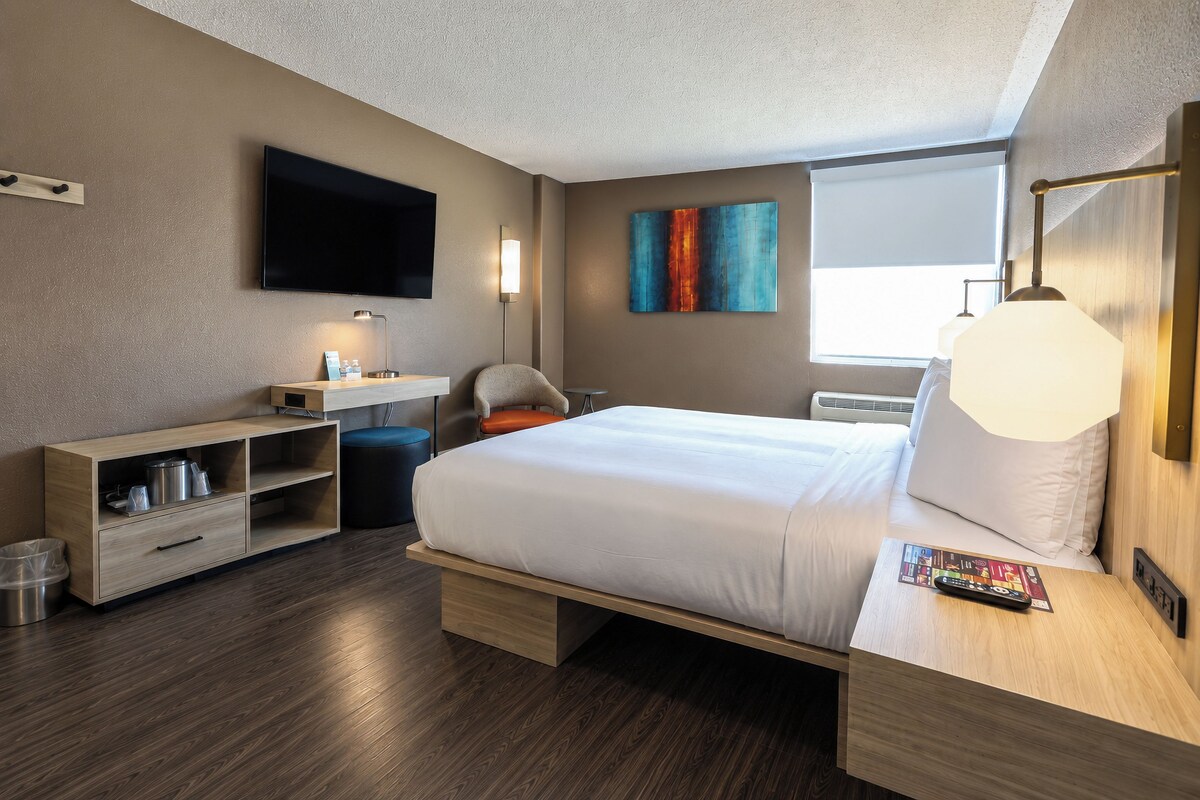 A hotel room with a bed, a desk, a chair, a TV, a painting, and a window.
