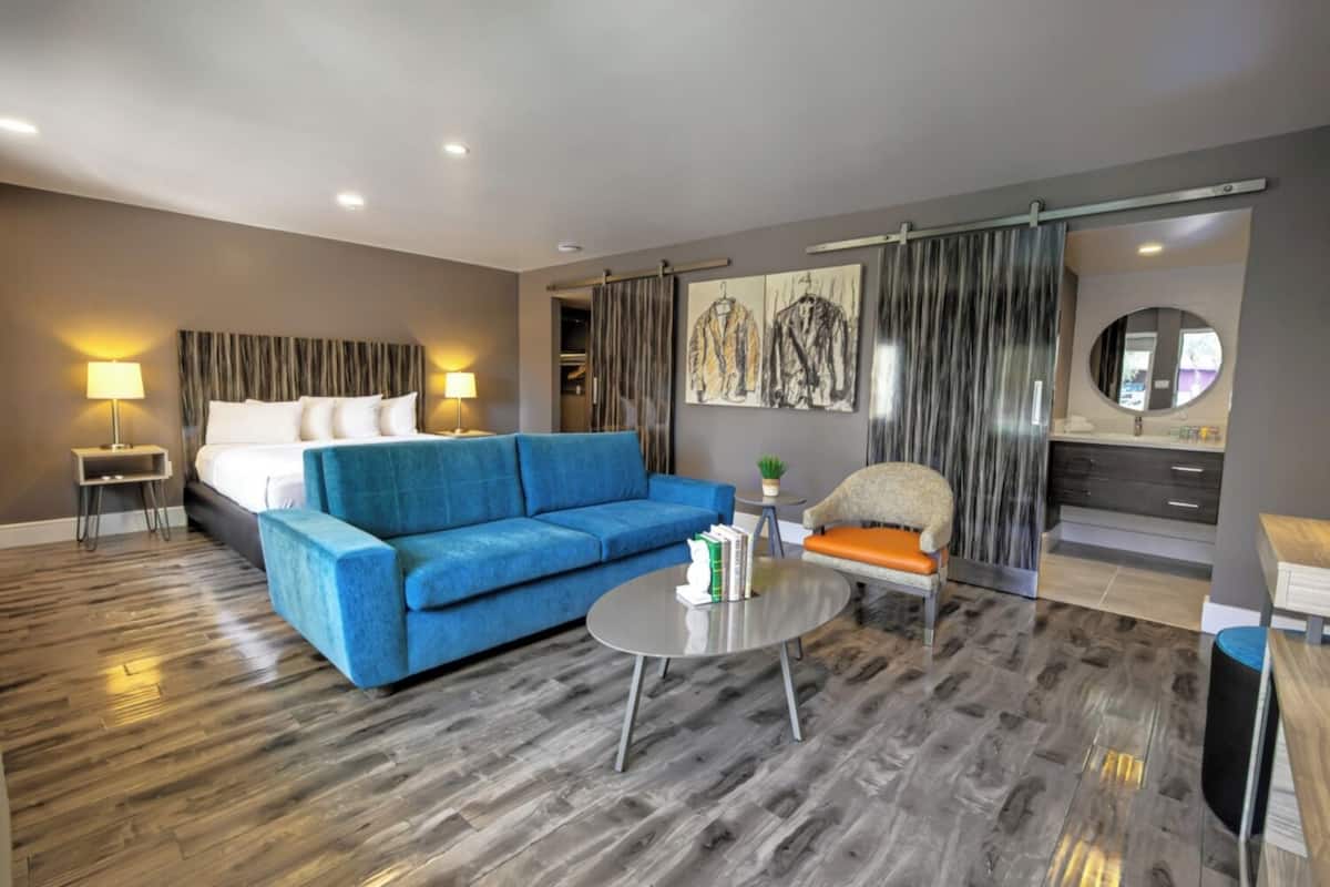 A modern hotel room with a large bed, a blue sofa, a round coffee table, a small armchair, and a bathroom with a vanity and mirror.