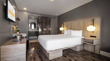 A modern hotel room with a large bed, bedside tables, a desk with a lamp, a mirror, and a TV on the wall.