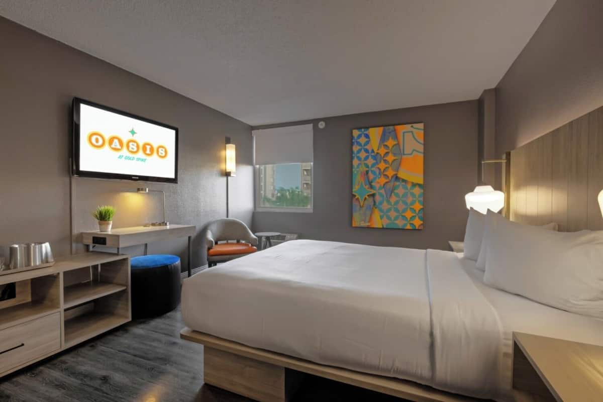 A modern hotel room with a large bed, a desk, a chair, and a TV displaying 'OASIS AT GOLD SPIKE'.