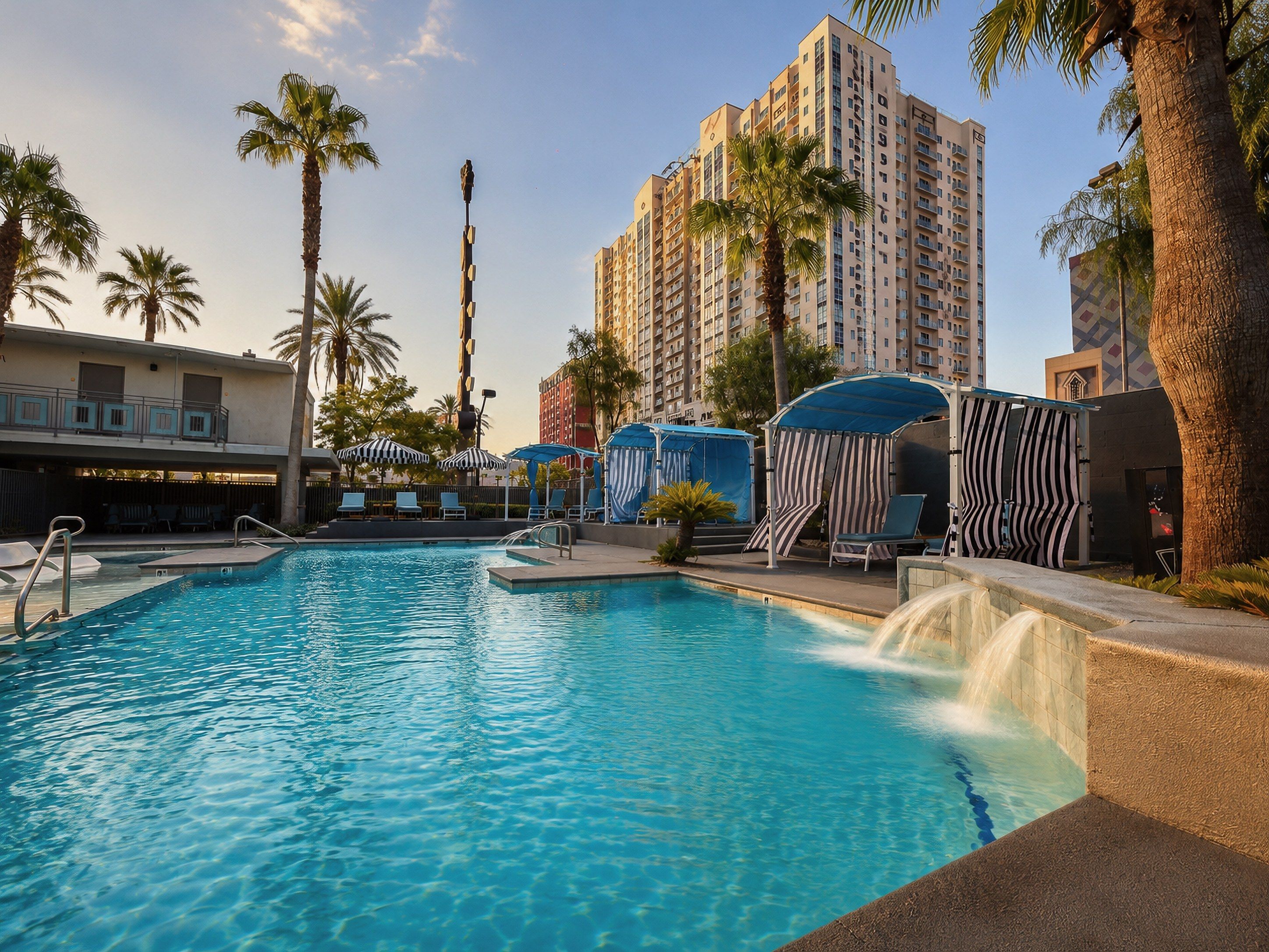 A swimming pool with lounge chairs and umbrellas, surrounded by palm trees and tall buildings.