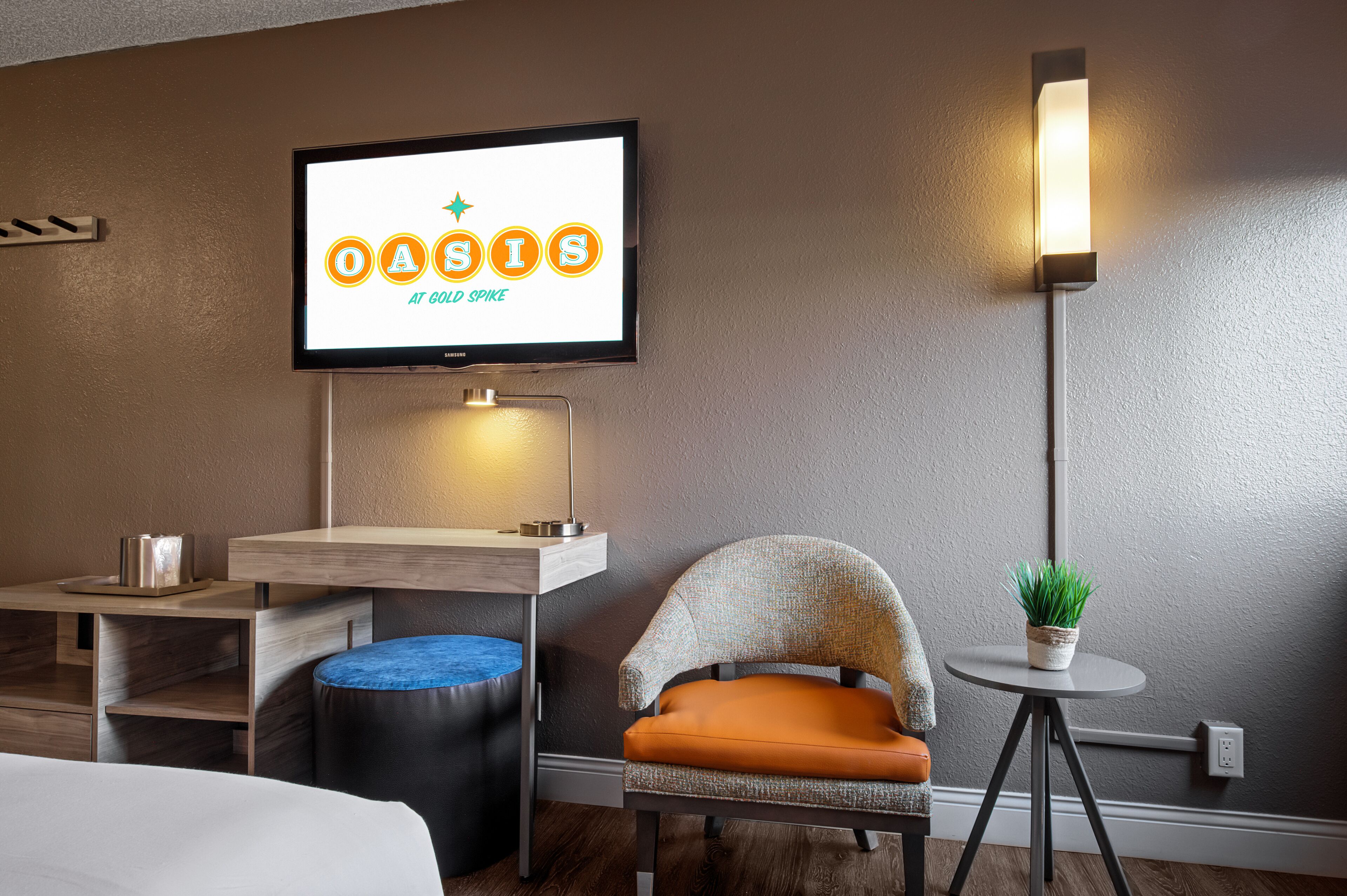 Hotelkamer met een televisie waarop 'OASIS at GOLD SPIKE' staat, een bureau met een lamp, een stoel met een oranje kussen, een klein rond tafeltje met een plant en een blauwe poef.