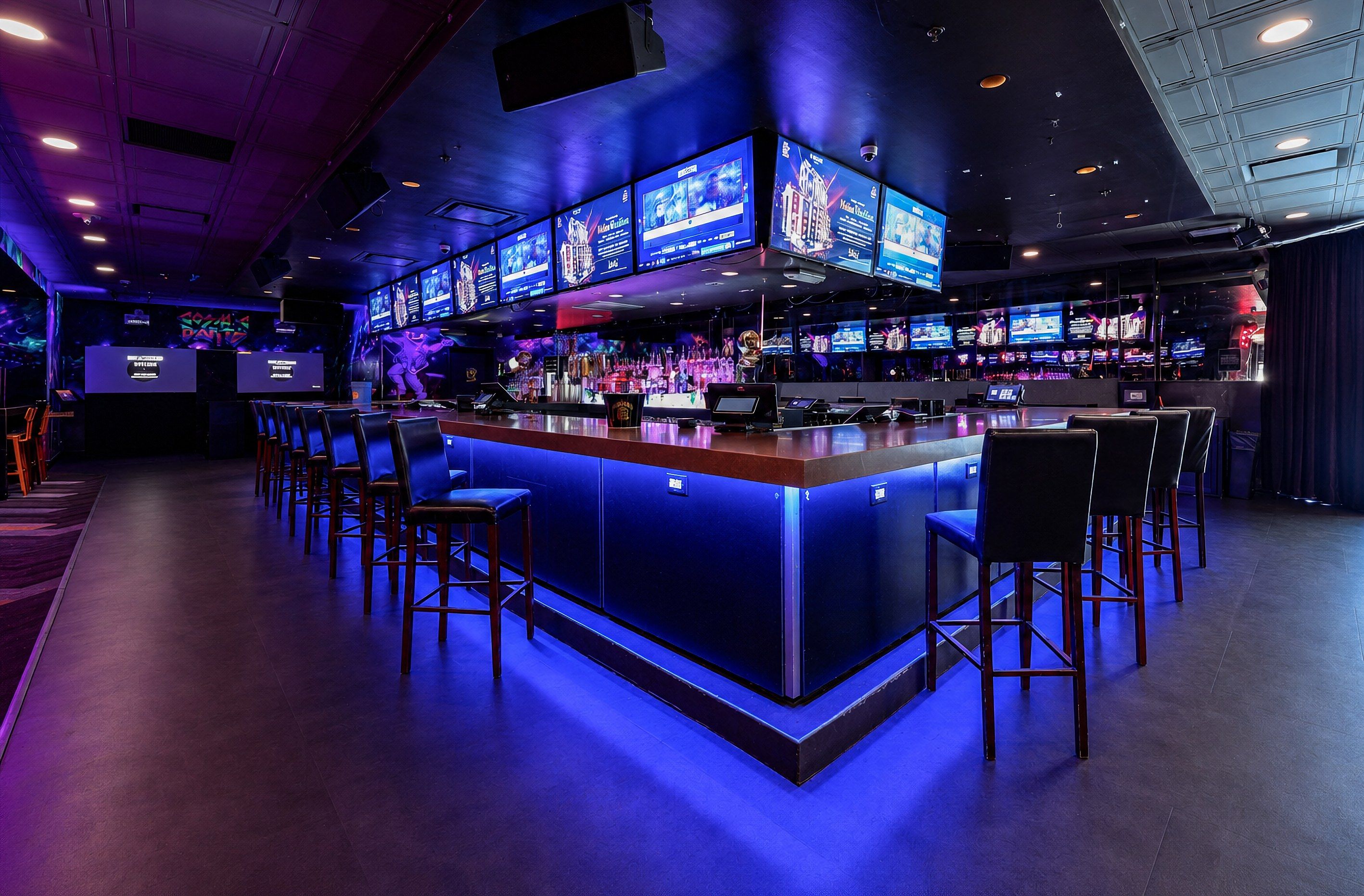 A bar area with multiple screens displaying content, bar stools, and a counter.
