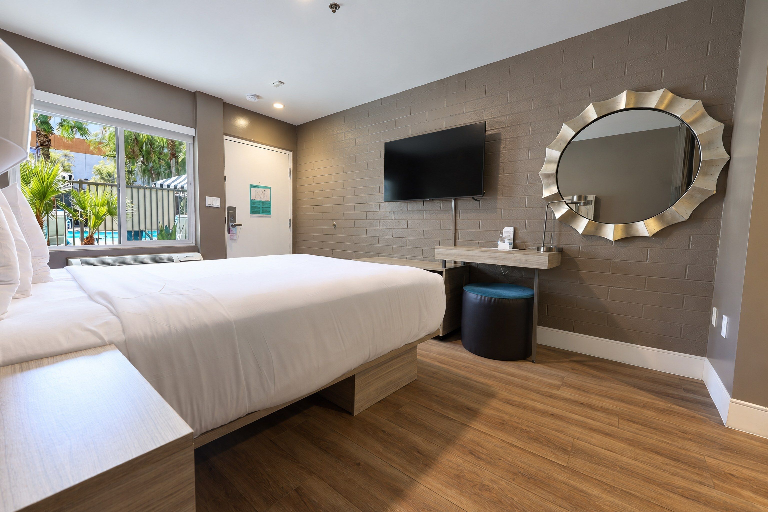 A modern hotel room with a bed, a TV, a round mirror, and a window with a view of palm trees.