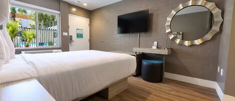 A modern hotel room with a bed, a TV, a round mirror, and a window with a view of palm trees.