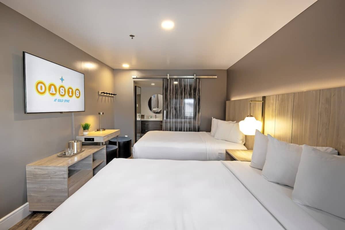 A hotel room with two beds, a desk, a mirror, and a TV displaying 'OASIS AT GOLD SPIKE'.