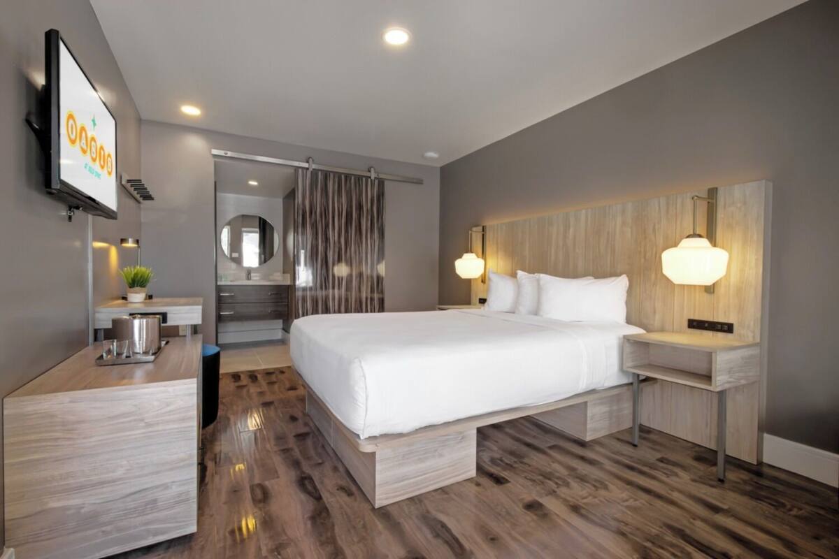 A modern hotel room with a large bed, bedside tables, a desk with a lamp, a mirror, and a TV displaying 'OAB18'.