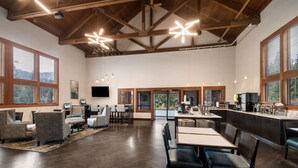 A spacious room with a high ceiling, wooden beams, and large windows. There are multiple seating areas with tables and chairs, and a kitchen area with modern appliances. - Best Western Mt. Hood Inn (Government Camp)