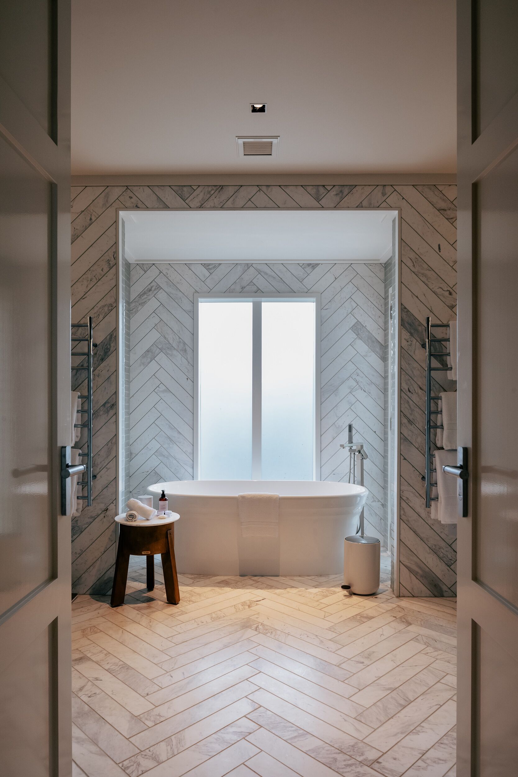 A modern bathroom with a herringbone-patterned wall, a freestanding bathtub, a small stool, and a towel rack.