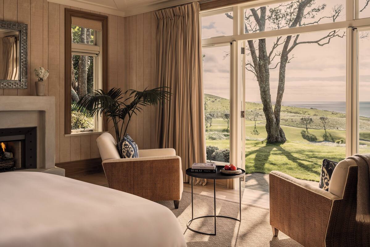 A spacious bedroom with a large bed, a sitting area with a chair and a small table, a fireplace, and a large window offering a scenic view of trees and hills.