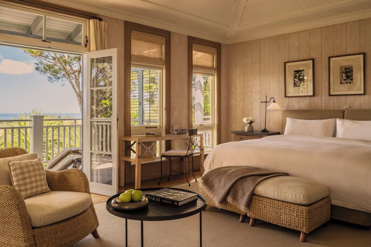 A bedroom with a large bed, a desk, and a chair. There is a balcony with a view of the ocean.