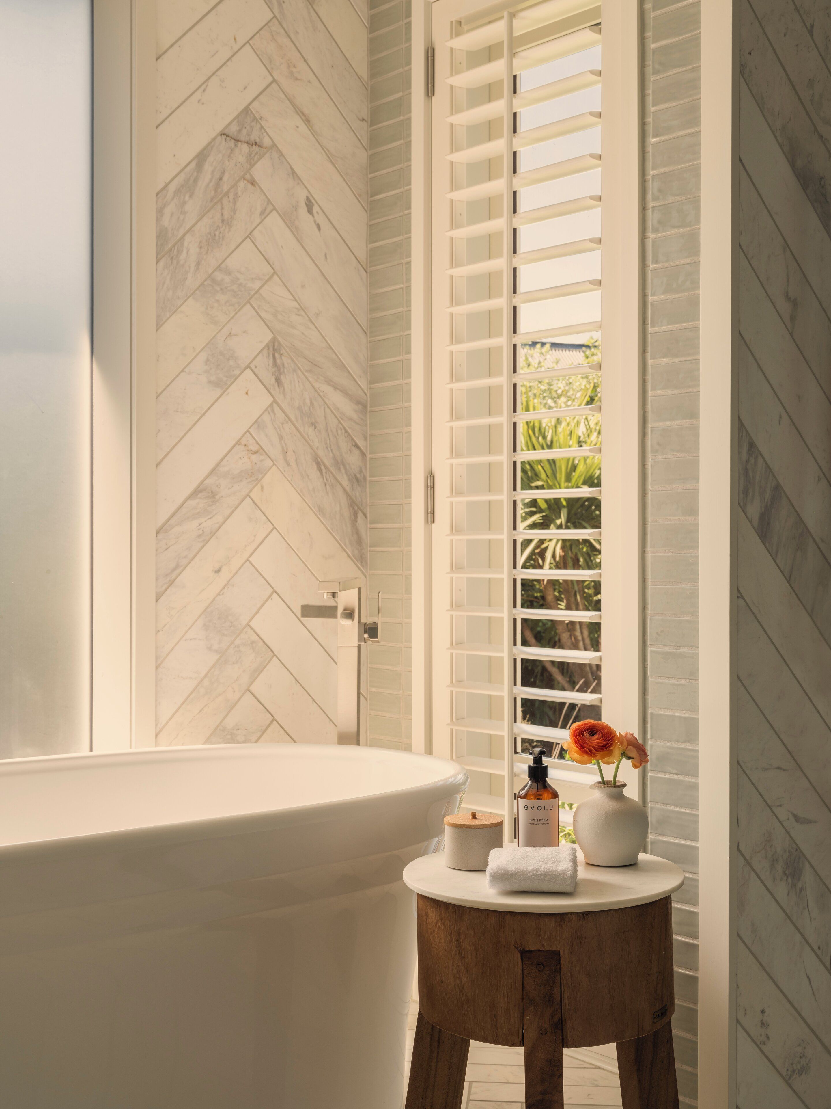 A bathroom with a herringbone-patterned floor, a freestanding bathtub, and a window with white shutters.
