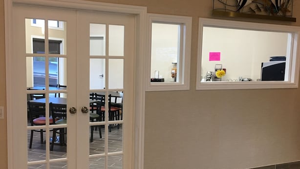 A spacious room with a wooden floor, a dining area visible through a glass door, and a small kitchen area with a microwave and a pink sign. - Bay Hill Inns & Suites, Neepawa (Neepawa)