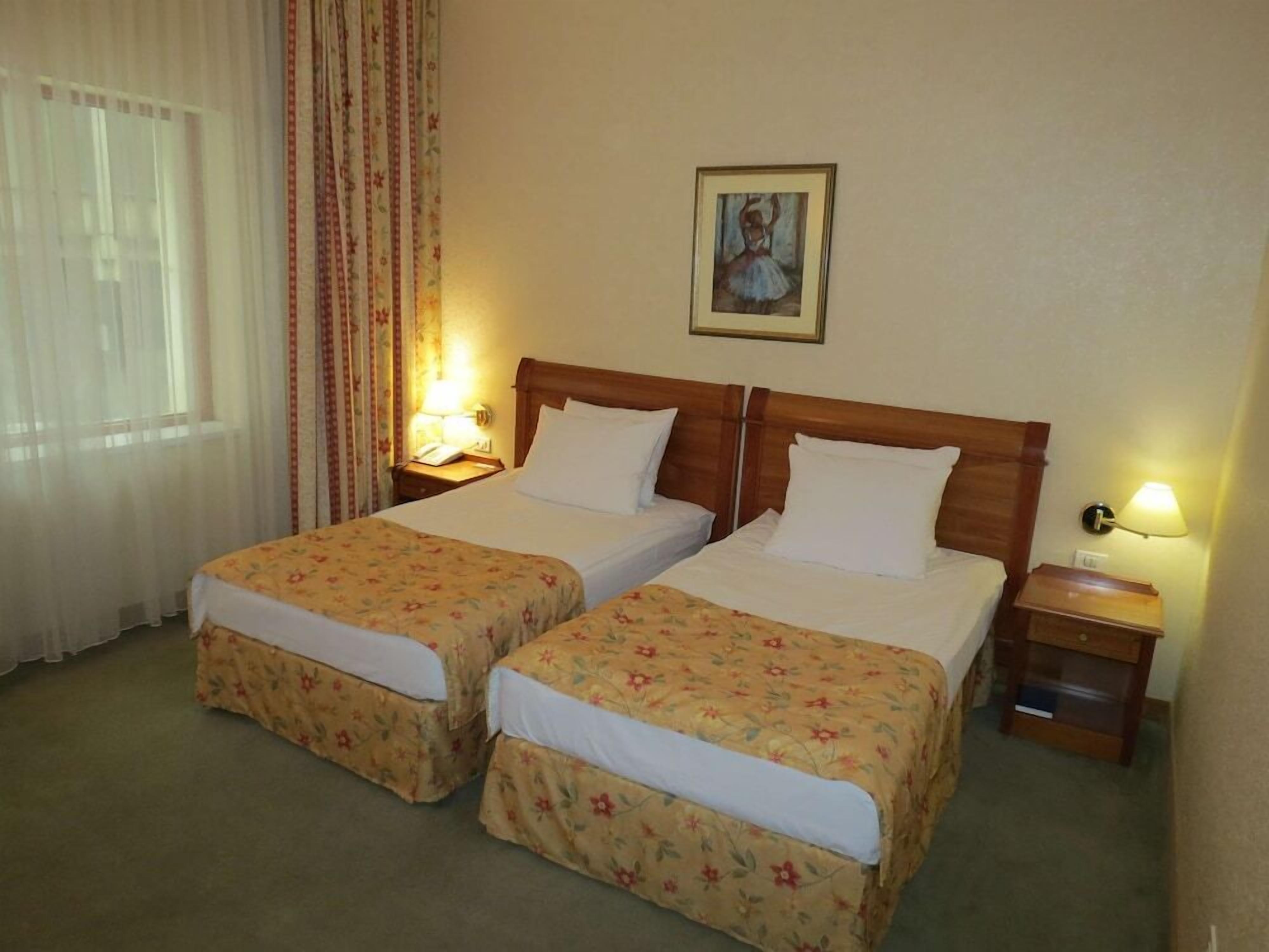 A hotel room with two single beds, a wooden headboard, a nightstand with a lamp, a framed picture on the wall, and a window with sheer curtains.