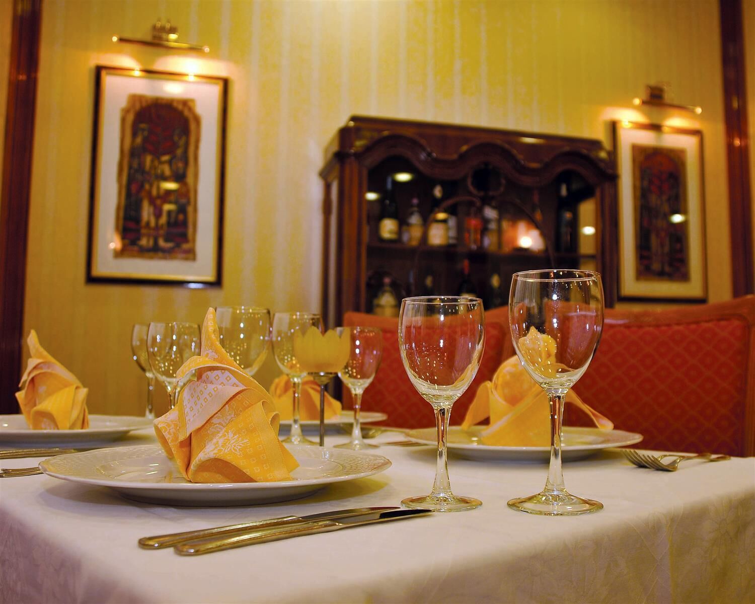 A dining table set with wine glasses and folded napkins, with a bar and framed pictures in the background.