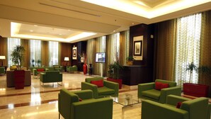A spacious lobby with green sofas, red cushions, and a glass coffee table. There are potted plants and framed pictures on the walls.