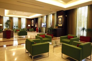 A spacious lobby with green sofas, red cushions, and a glass coffee table. There are potted plants and framed pictures on the walls.