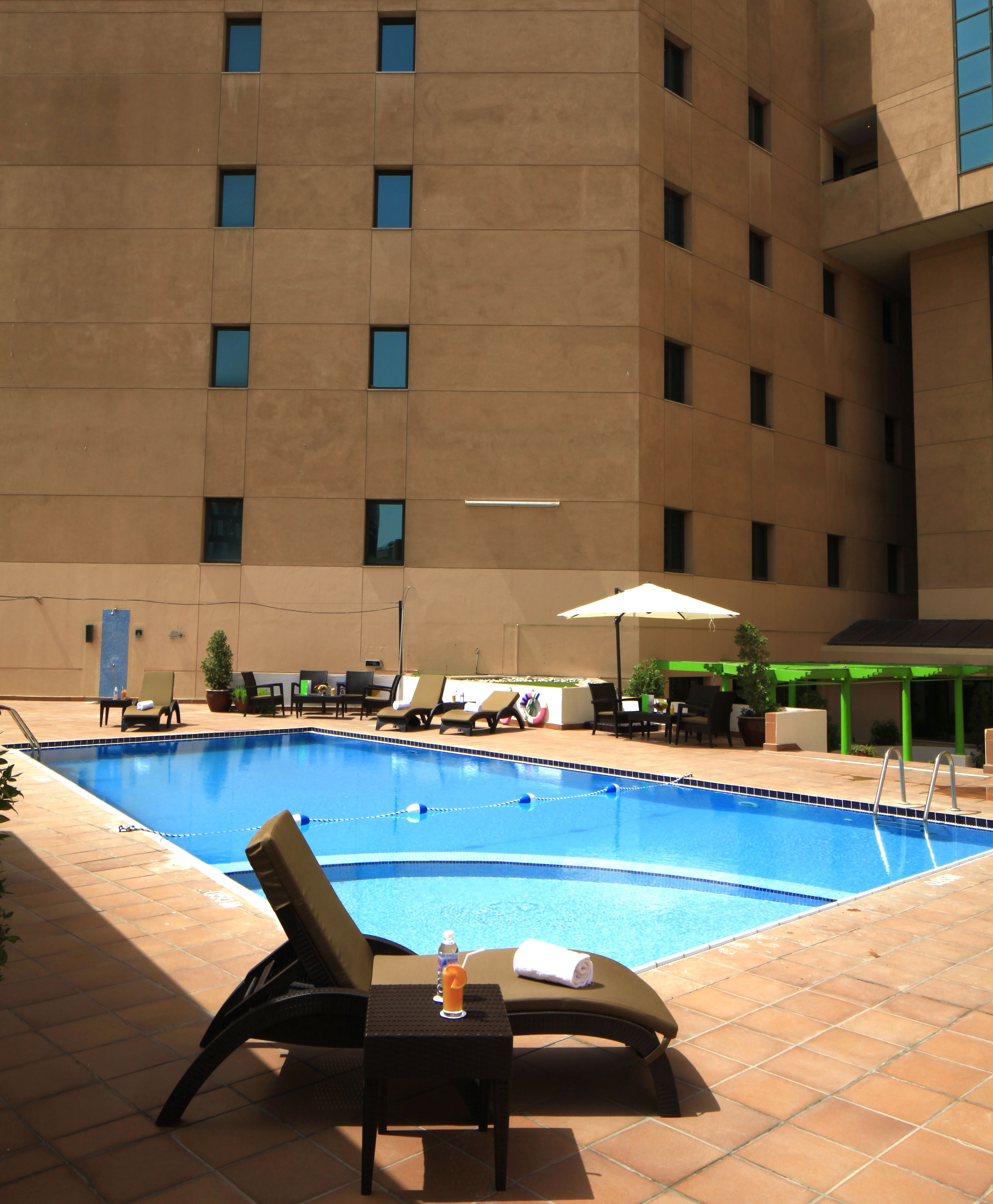 Piscina con sillas de descanso y sombrillas, rodeada por un edificio con numerosas ventanas.