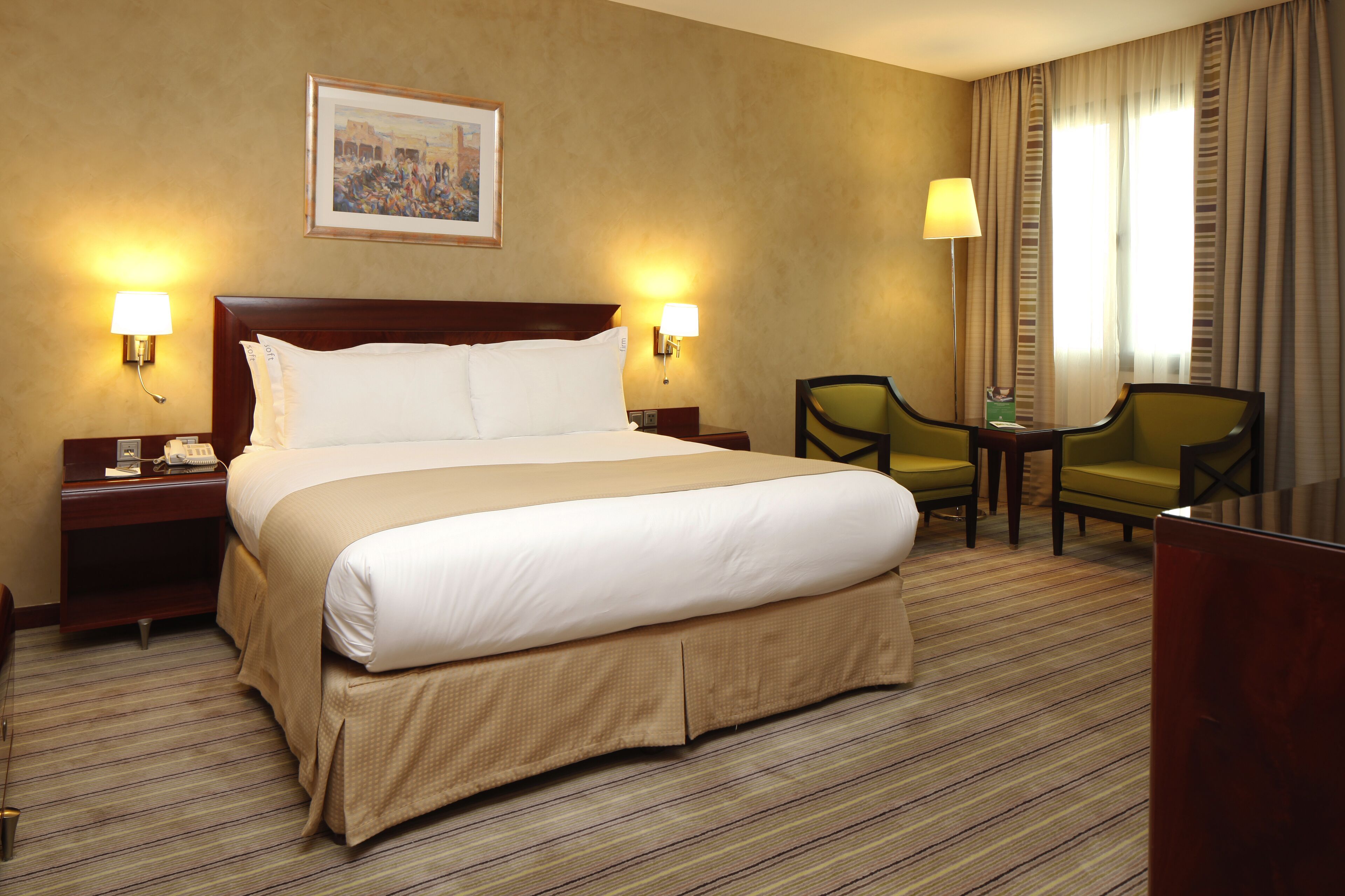 A hotel room with a large bed, two bedside lamps, a nightstand, a desk with a chair, and a framed picture on the wall.