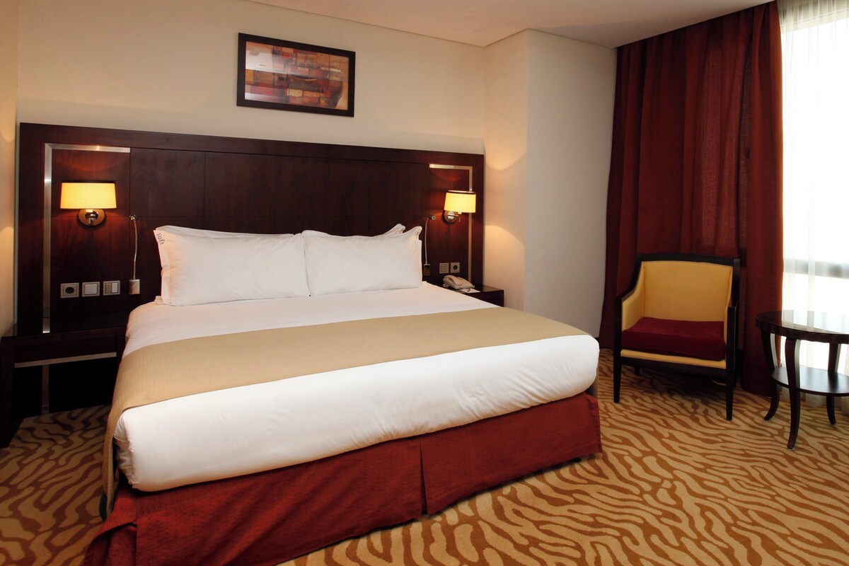 A hotel room with a large bed, two bedside tables with lamps, a chair, a painting on the wall, and a window with curtains.