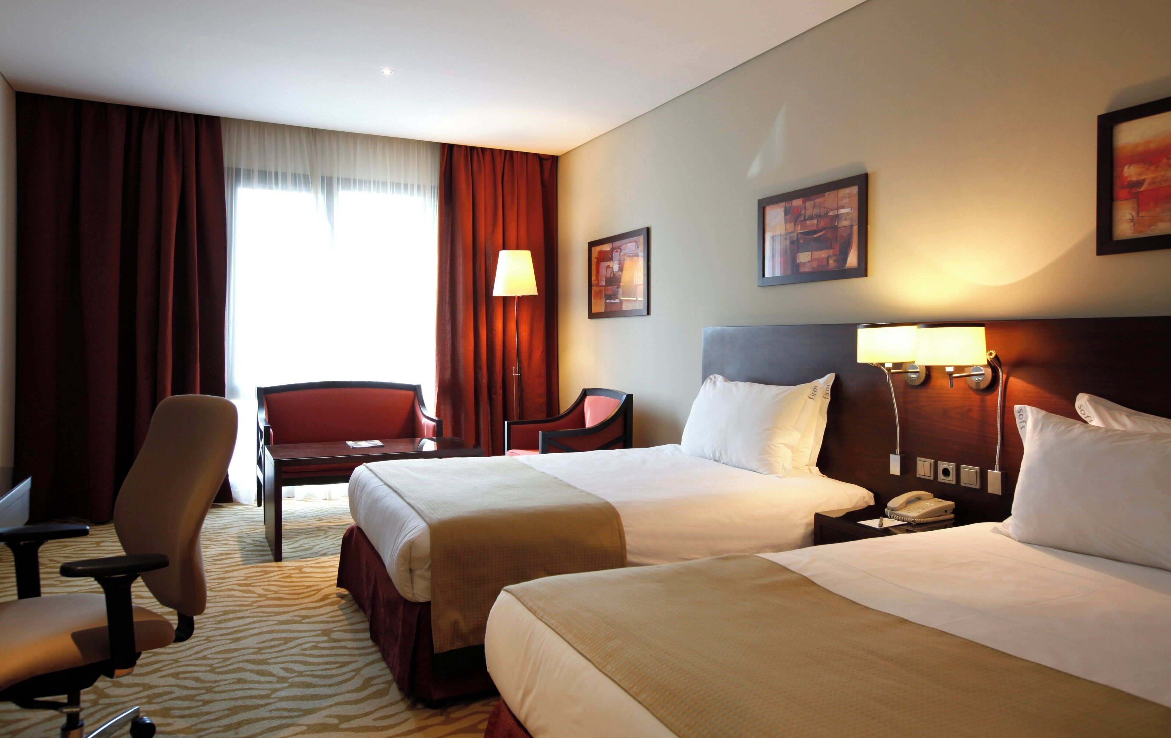 A hotel room with two beds, a desk, a chair, and a lamp.