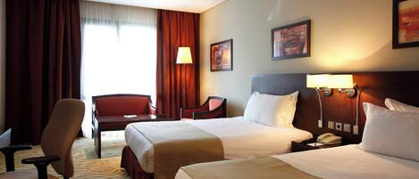 A hotel room with two beds, a desk, a chair, and a lamp.