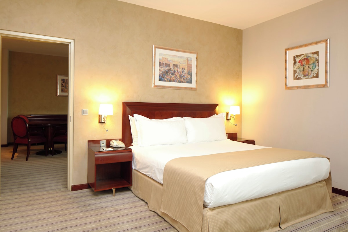 A hotel room with a large bed, two bedside lamps, a desk with a chair, and two framed pictures on the wall.