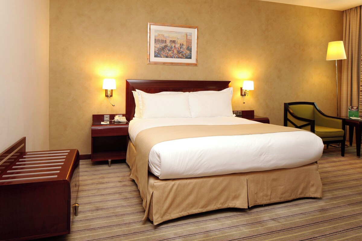 A hotel room with a large bed, two bedside tables with lamps, a chair, and a painting on the wall.