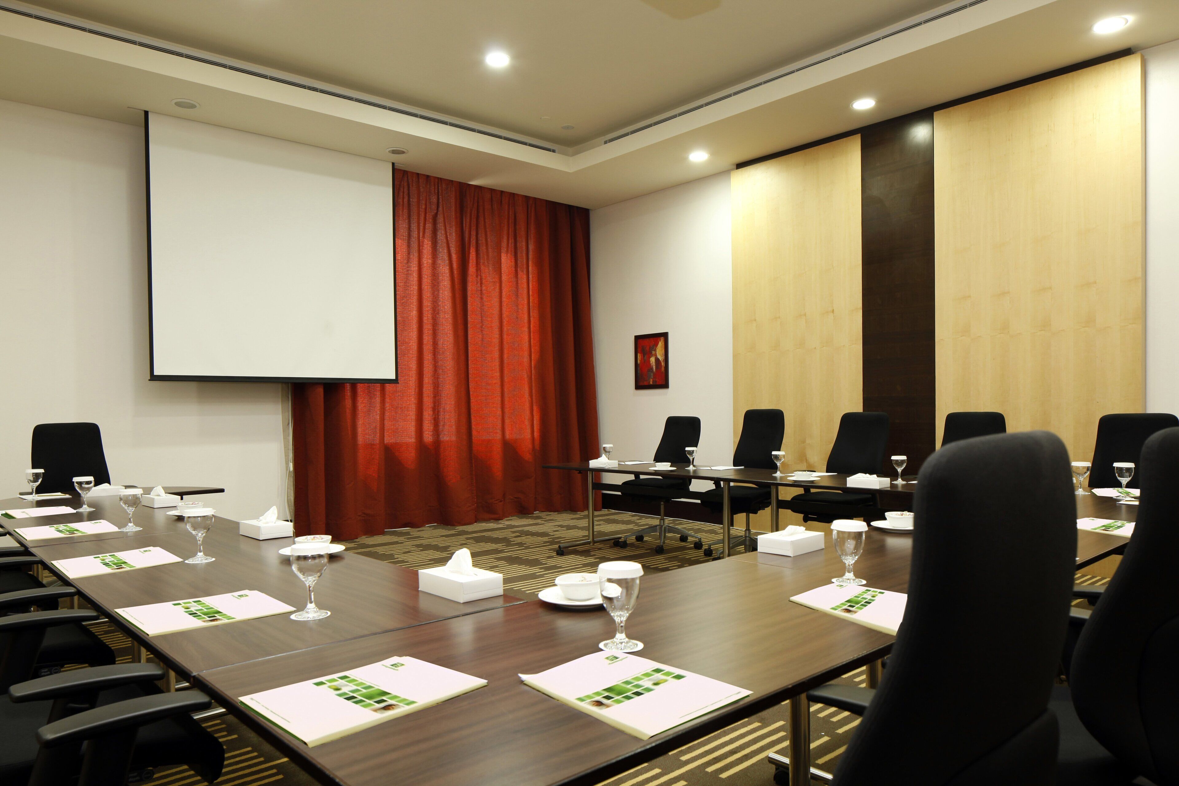 Un salón de conferencias con una larga mesa, sillas, una pantalla de proyector y una cortina roja.