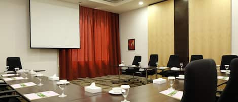 Un salón de conferencias con una larga mesa, sillas, una pantalla de proyector y una cortina roja.