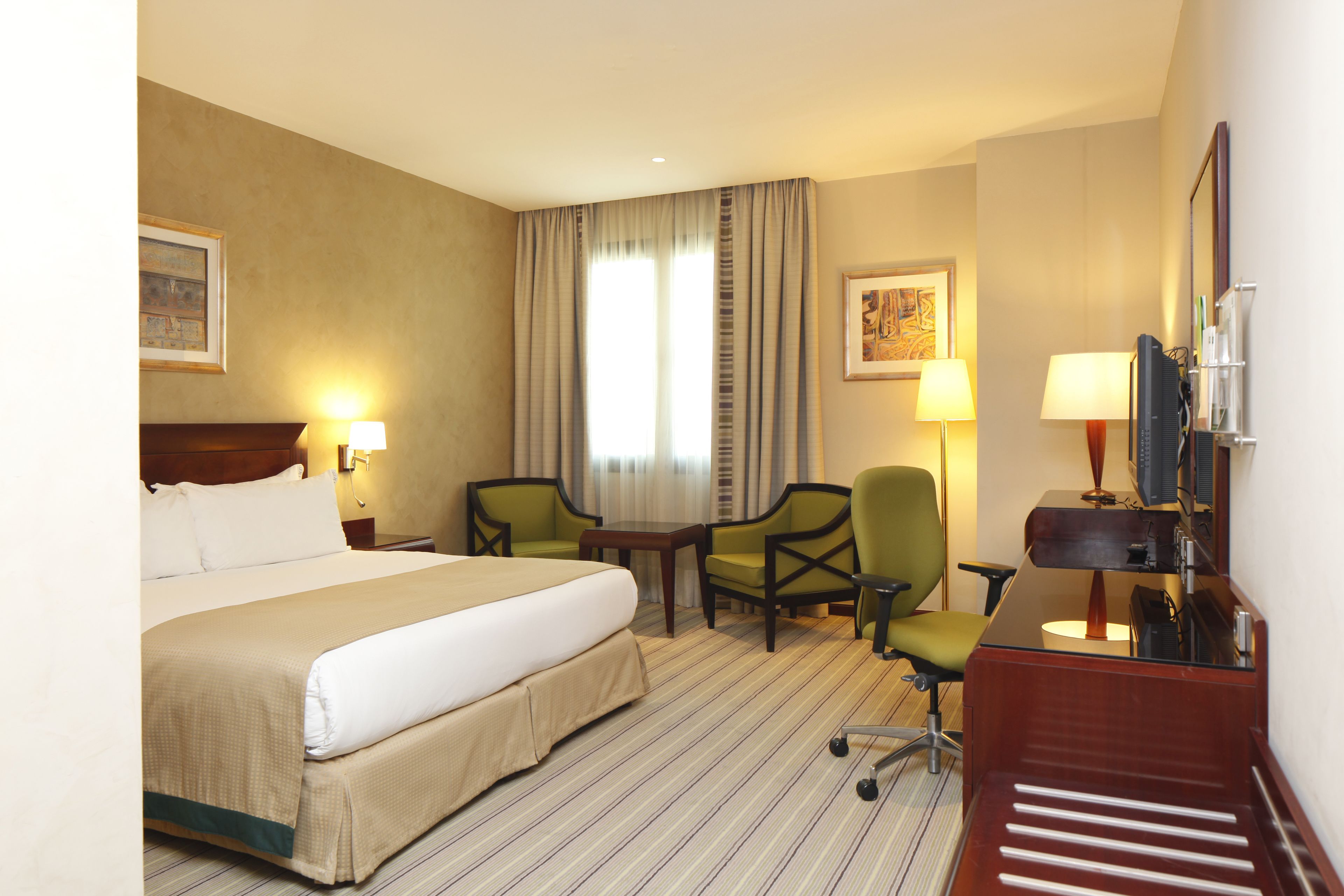 A hotel room with a large bed, two chairs, a desk with a computer, and a TV.