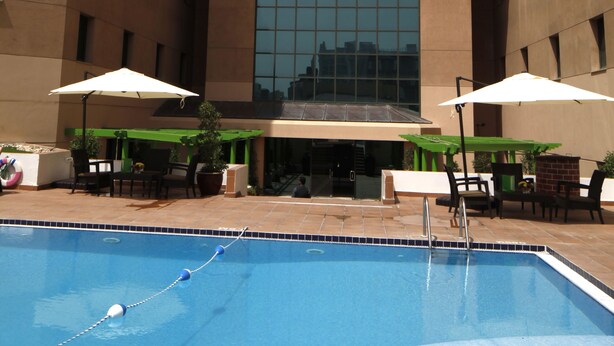 A swimming pool with lounge chairs and umbrellas, surrounded by a building with a large glass window and a clock.