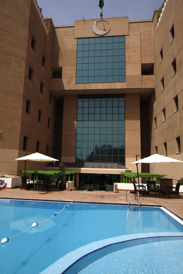 A swimming pool with lounge chairs and umbrellas, surrounded by a building with a large glass window and a clock.