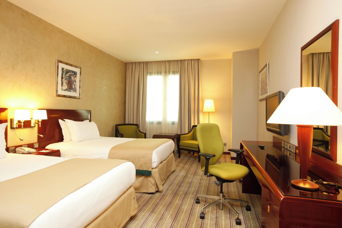 A hotel room with two beds, a desk, a chair, and a TV.