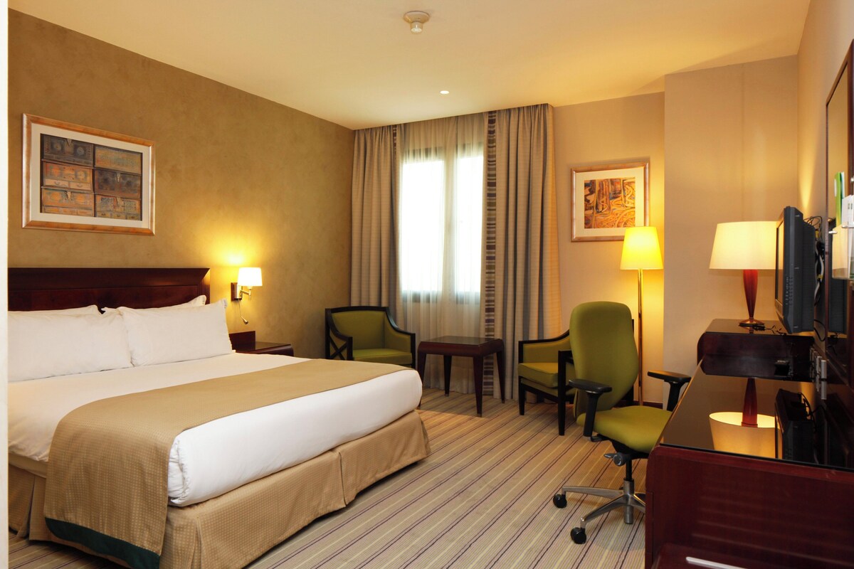A hotel room with a large bed, two chairs, a desk with a computer, and two lamps.