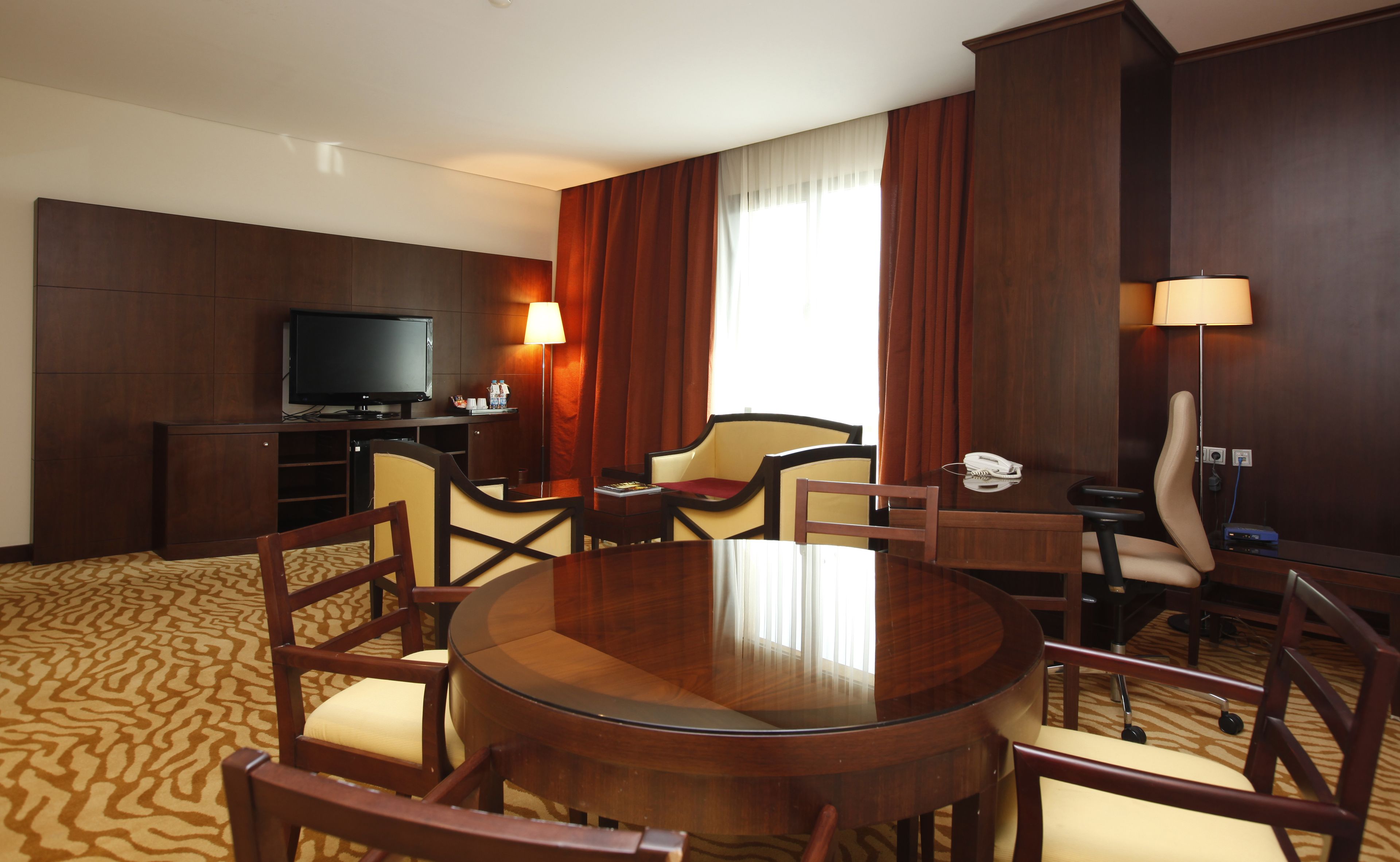 A hotel room with a round table, four chairs, a television, a desk with a lamp, and a large window with curtains.
