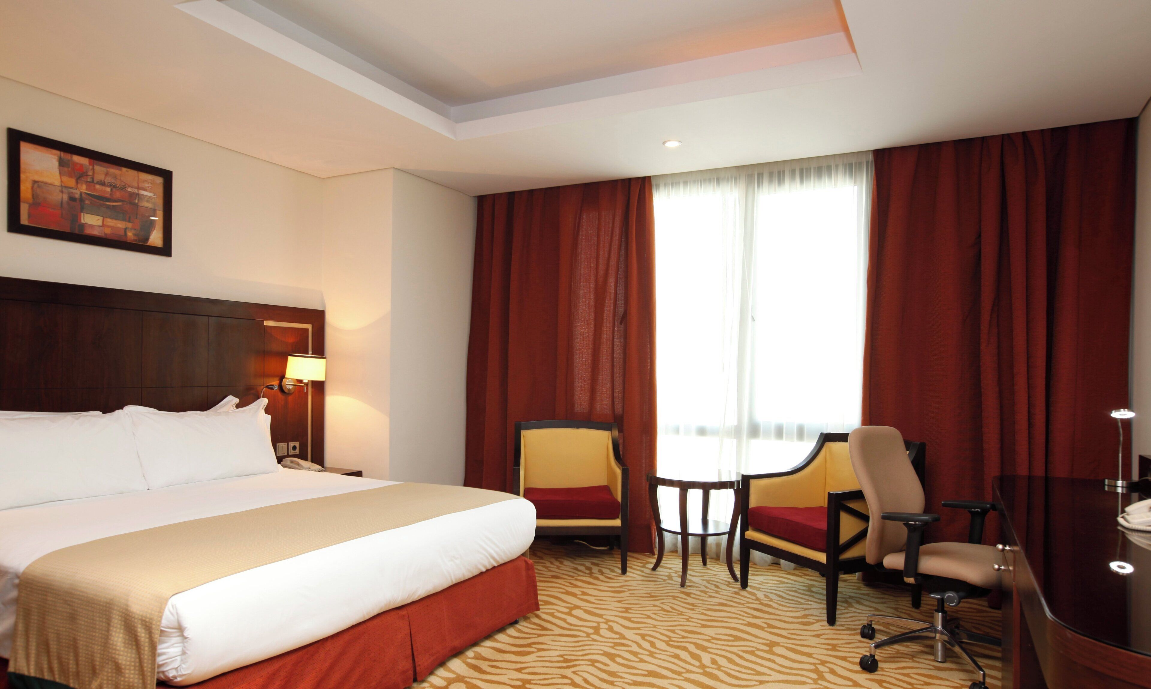 A hotel room with a large bed, a desk, two chairs, a small table, and a window with curtains.