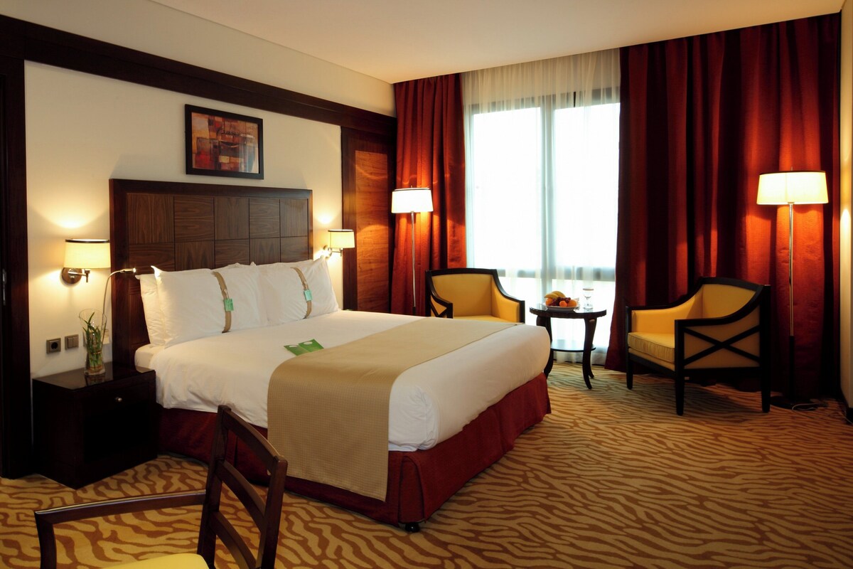 A hotel room with a large bed, two chairs, a small table with fruit and a glass, and a window with curtains.
