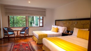 Habitación de hotel con dos camas, una mesita con sillas y vistas al exterior con vegetación.