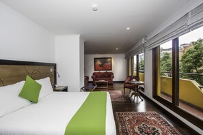 A hotel room with a large bed, a green accent pillow, a small table, a sofa, and a large window offering a view of the outside. - Hotel Emaus Bogotá (Bogotá)