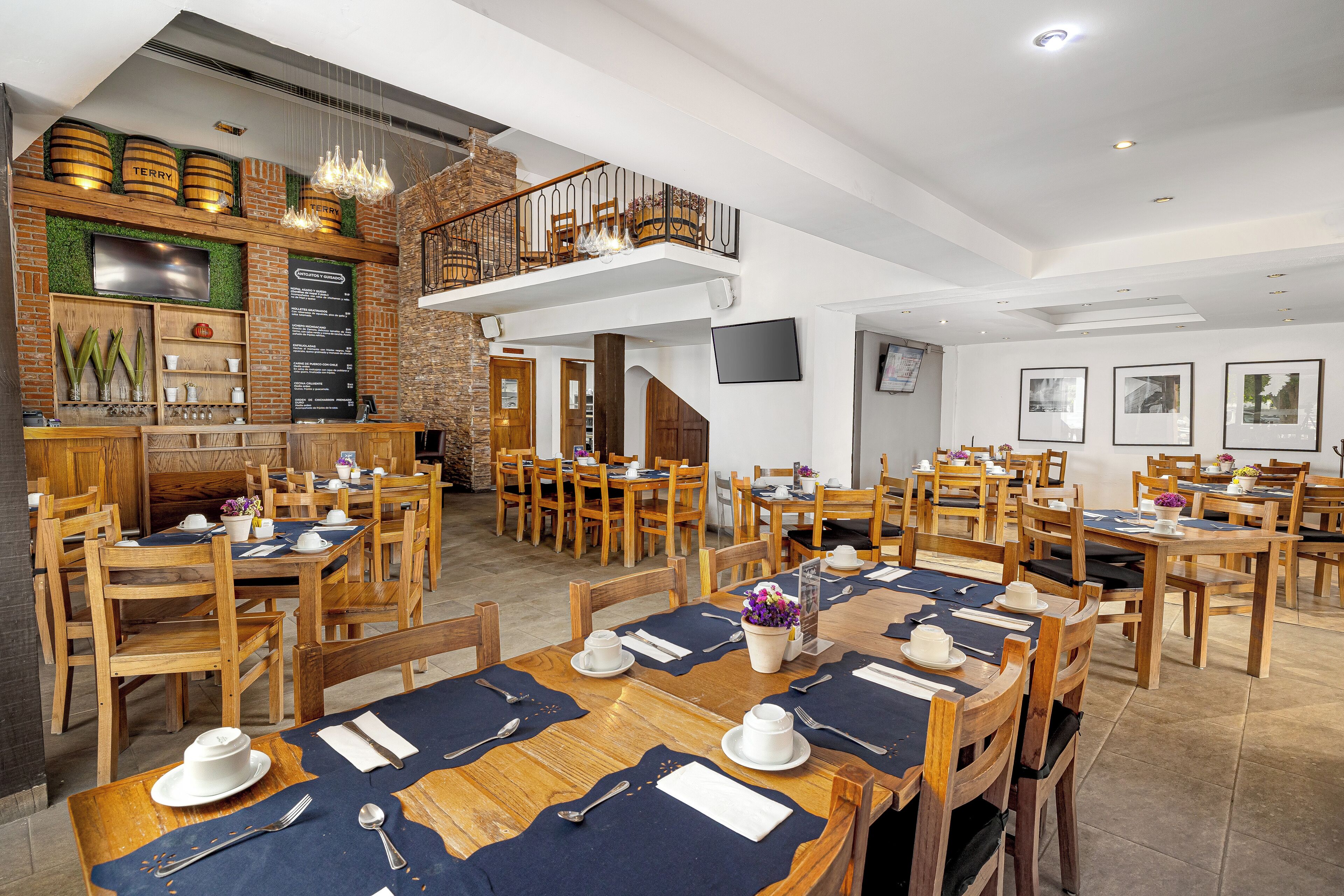 A restaurant with wooden tables and chairs, a brick wall with a TV, and a staircase leading to a balcony.