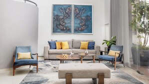 Een ruime woonkamer met een hoge plafond, een grijze bank met gele kussens, een blauwe fauteuil, een houten salontafel en abstracte schilderijen aan de muur.