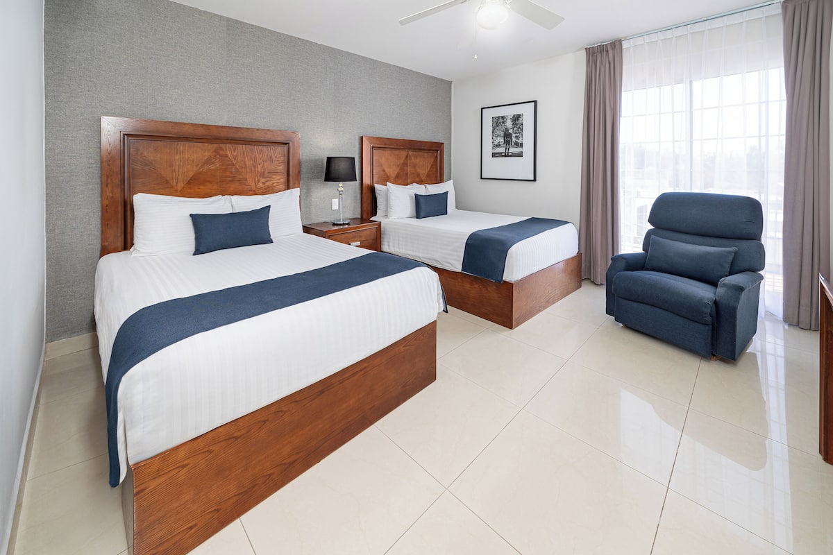 A hotel room with two wooden beds, a blue armchair, a nightstand with a lamp, and a framed picture on the wall.