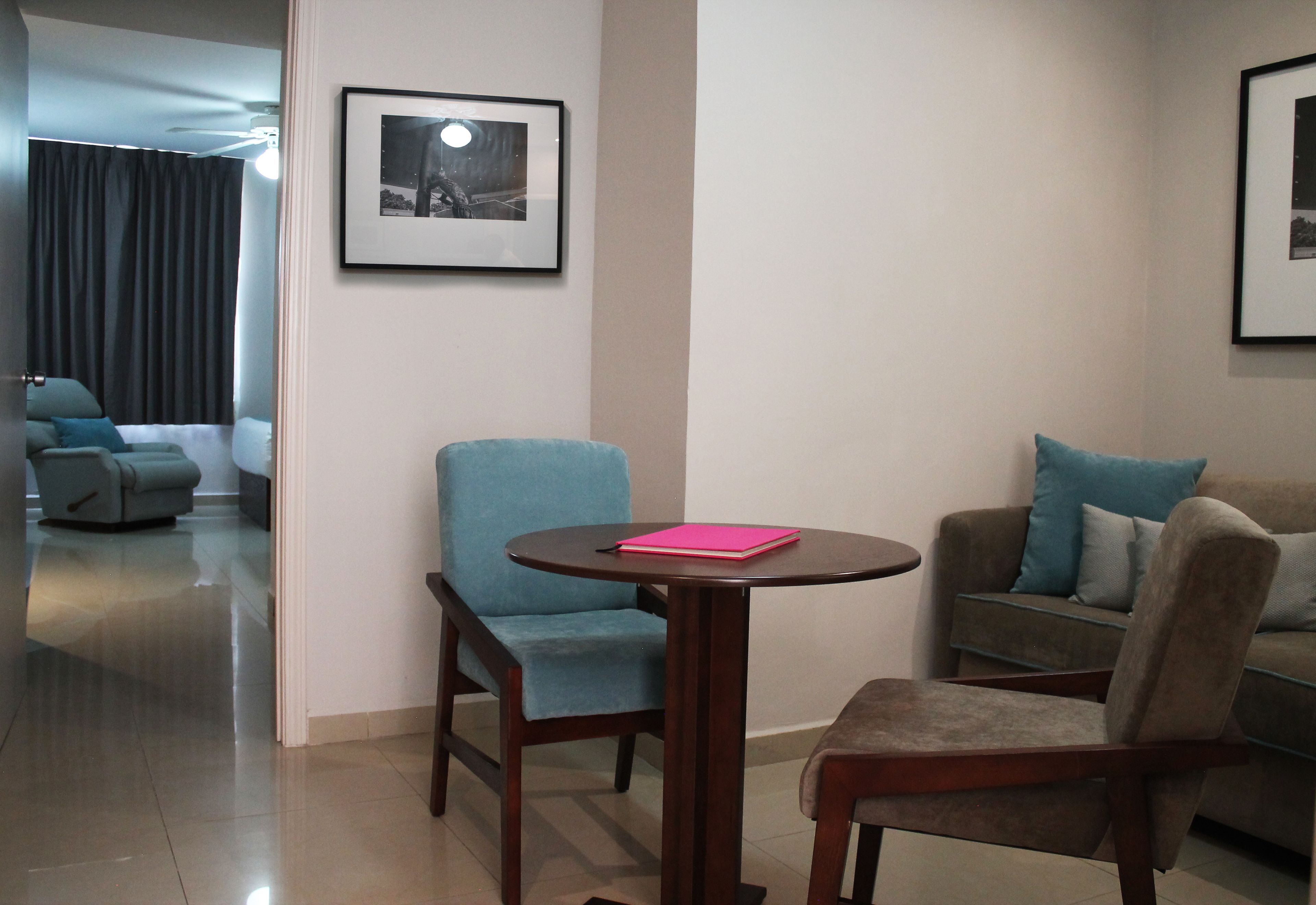 Una sala de estar moderna con una mesa redonda de madera, dos sillones y un sofá. Se observa un cuadro en la pared y un ventilador de techo.