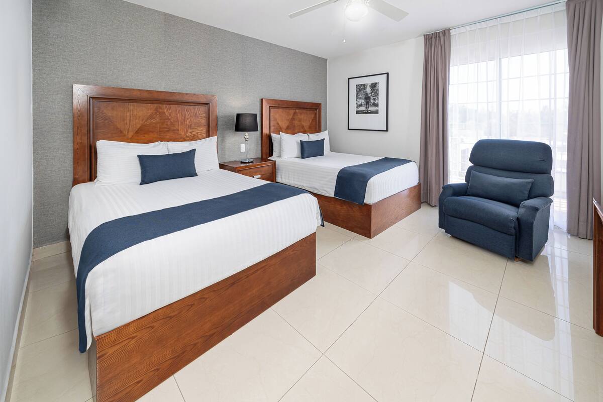 A hotel room with two wooden beds, a blue armchair, a nightstand with a lamp, and a framed picture on the wall.