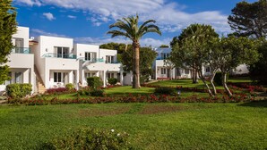 A modern hotel with white exterior walls, multiple balconies, and a well-maintained garden with red flowers and a palm tree.