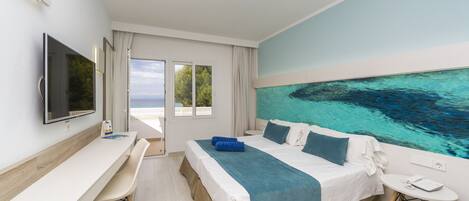 A modern hotel room with a large bed, a desk, a television, and a view of the sea through the window.