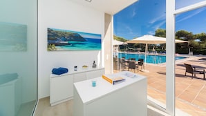 Un baño moderno con un ventanal grande, un cuadro de una escena costera y vistas a una zona de piscina exterior.